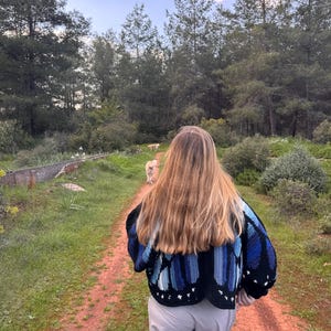 May include: A person walking on a dirt path in a forest, wearing a black and blue crochet cardigan. The cardigan has a butterfly wing design. The person is wearing light gray pants. A dog is walking ahead on the path.