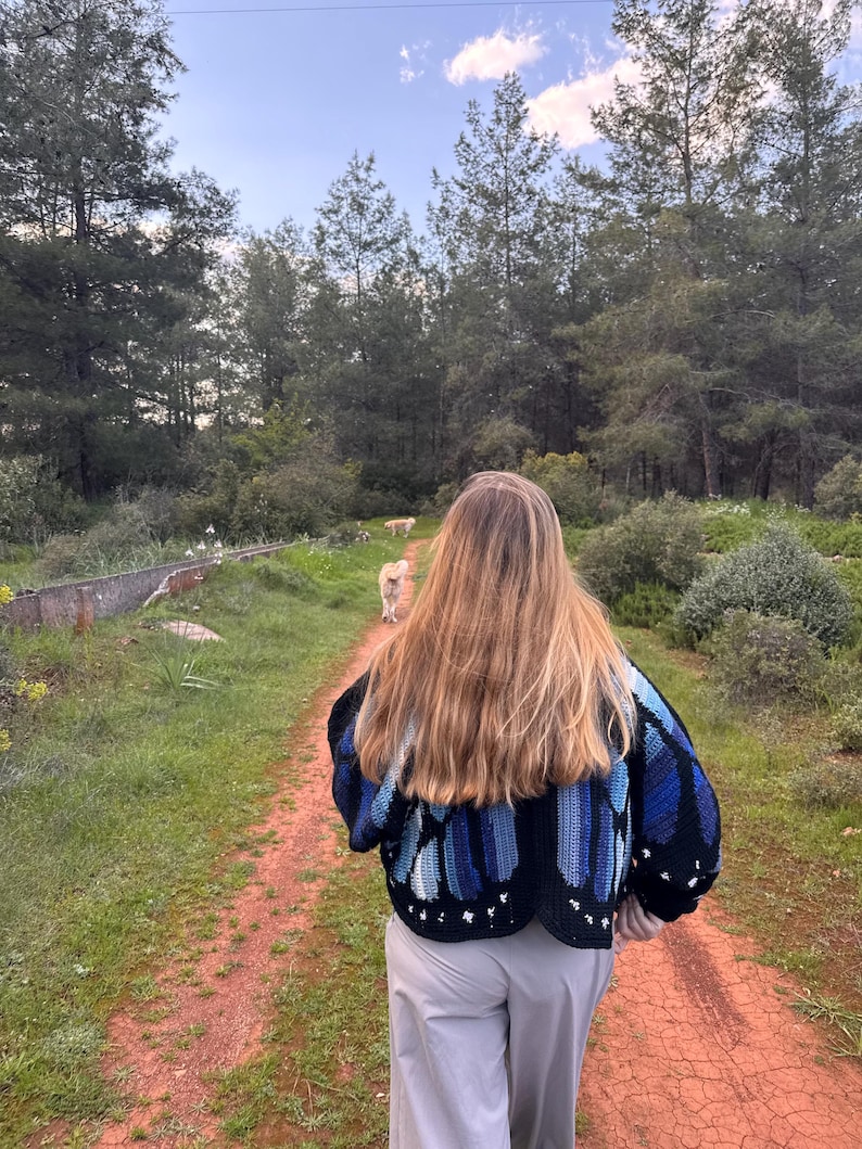 May include: A person walking on a dirt path in a forest, wearing a black and blue crochet cardigan. The cardigan has a butterfly wing design. The person is wearing light gray pants. A dog is walking ahead on the path.