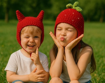 May include: Two children wearing red knitted hats. One hat is shaped like a devil with horns, and the other is a strawberry. The children are smiling and making faces while sitting on green grass.