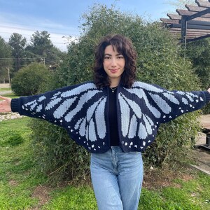 May include: A dark navy blue and light blue crochet cardigan featuring a butterfly wing pattern across the back and sleeves. The garment has an open front design with ribbed cuffs and a solid dark navy blue border along the neckline and hem.