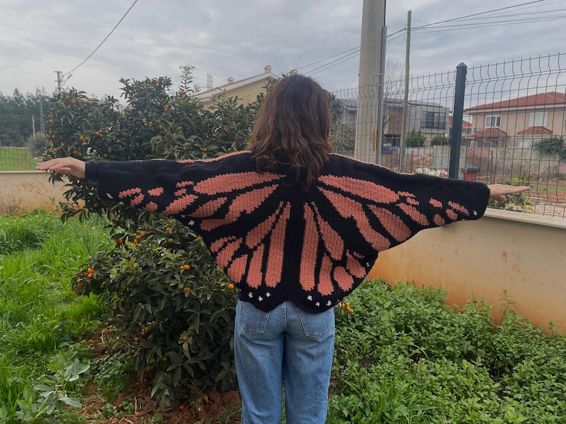 May include: A black and peach-colored crochet cardigan featuring a large butterfly wing pattern that spans across the back and sleeves. The garment has long sleeves and a scalloped hemline, worn over blue denim jeans in an outdoor setting.