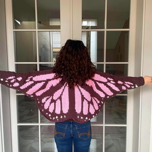 May include: A dark brown crochet cardigan featuring a pink butterfly wing pattern across the back and sleeves. The garment has long sleeves and a wide, open-wing design that extends across the shoulders and arms, with small white accents.