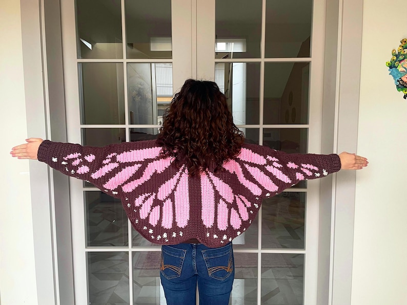 May include: A dark brown crochet cardigan featuring a pink butterfly wing pattern across the back and sleeves. The garment has long sleeves and a wide, open-wing design that extends across the shoulders and arms, with small white accents.
