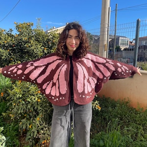 May include: A crochet cardigan featuring a butterfly wing pattern in dark brown and light pink. The garment has an open front, wide sleeves, and a rounded hemline, with the wing design extending across the back and front panels of the piece.