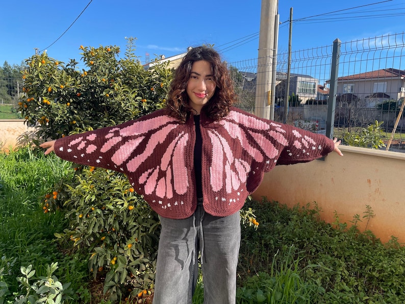 May include: A crochet cardigan featuring a butterfly wing pattern in dark brown and light pink. The garment has an open front, wide sleeves, and a rounded hemline, with the wing design extending across the back and front panels of the piece.