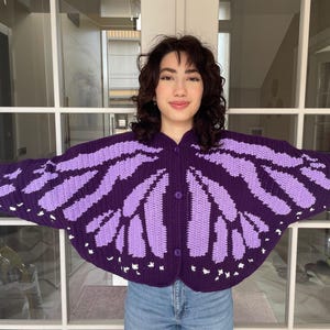 May include: A dark purple and light purple crocheted cardigan featuring a butterfly wing pattern across the front and sleeves. The garment has a rounded hem, long sleeves, and three matching dark purple buttons down the center front closure.