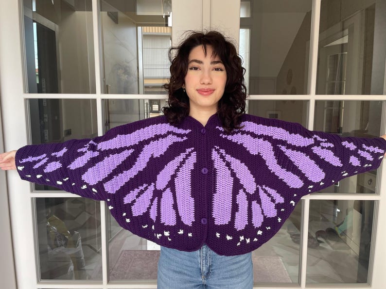May include: A dark purple and light purple crocheted cardigan featuring a butterfly wing pattern across the front and sleeves. The garment has a rounded hem, long sleeves, and three matching dark purple buttons down the center front closure.