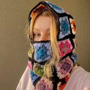Puede incluir: Una colorida capucha de ganchillo con una cobertura facial. La capucha presenta un diseño de patchwork con cuadrados en varios colores, incluyendo azul, rosa, verde y morado, delineados en negro. La capucha está diseñada para cubrir la cabeza y el cuello.