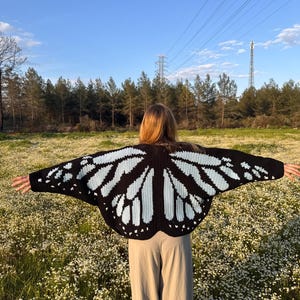 May include: A black and light blue crochet cardigan featuring a butterfly wing pattern across the back and sleeves. The garment is worn with light beige trousers, showcasing the intricate stitch work and the symmetrical wing design.
