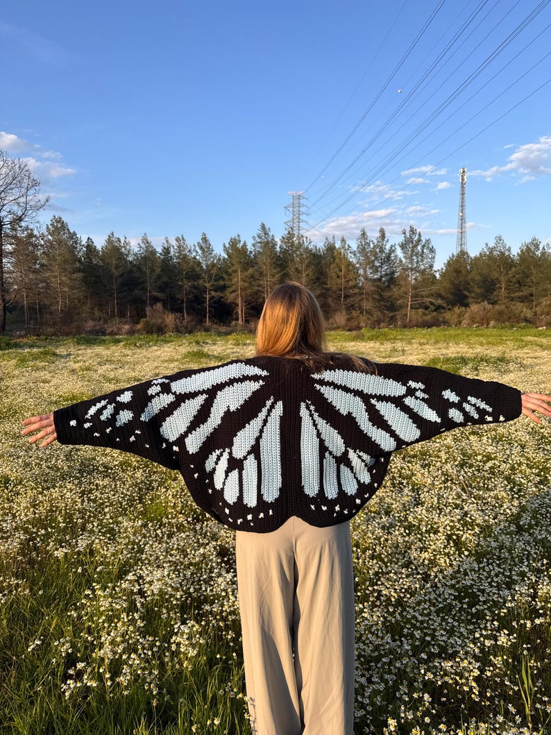 May include: A black and light blue crochet cardigan featuring a butterfly wing pattern across the back and sleeves. The garment is worn with light beige trousers, showcasing the intricate stitch work and the symmetrical wing design.