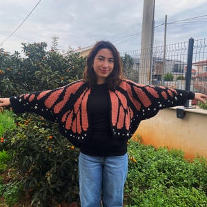 May include: A crochet cardigan featuring a black and orange monarch butterfly wing pattern. The garment has long sleeves, a wide open front, and small white accents along the black borders, mimicking the appearance of butterfly wing markings.