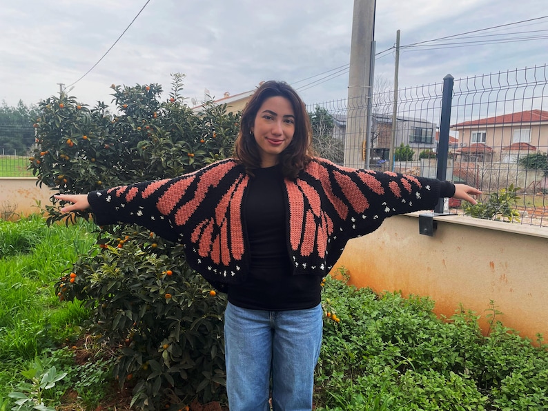 May include: A crochet cardigan featuring a black and orange monarch butterfly wing pattern. The garment has long sleeves, a wide open front, and small white accents along the black borders, mimicking the appearance of butterfly wing markings.