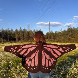 May include: A brown and pink butterfly wing-shaped crochet cardigan. The cardigan features a dark brown base with pink wing designs and white accents. The person is wearing blue jeans and standing in a field of white flowers.
