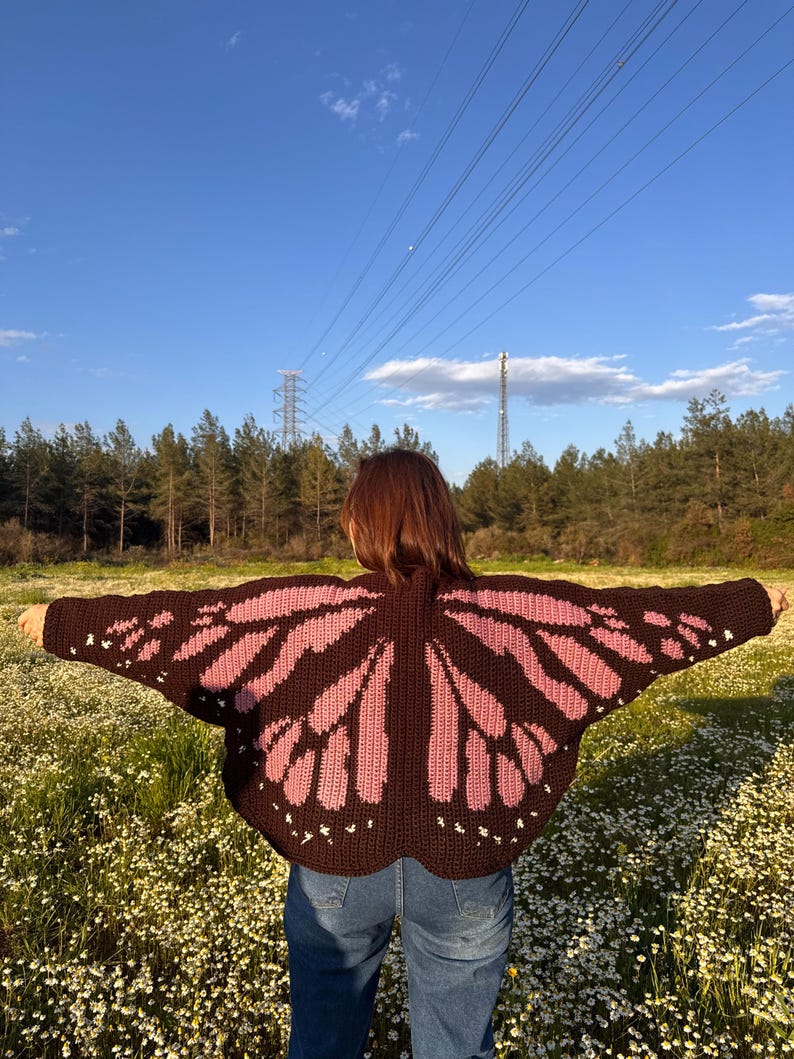 May include: A brown and pink butterfly wing-shaped crochet cardigan. The cardigan features a dark brown base with pink wing designs and white accents. The person is wearing blue jeans and standing in a field of white flowers.