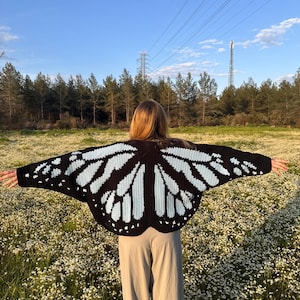 May include: A black and light blue butterfly-wing-shaped crochet sweater. The sweater is worn over a pair of tan pants. The background features a field of white flowers, trees, and a blue sky.