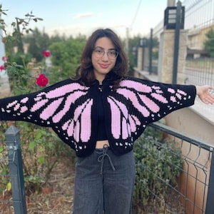 May include: A black crochet cardigan featuring a pink butterfly wing pattern on the sleeves and back. The garment has an open front design with wide, wing-like sleeves that extend outward, showcasing a detailed pink and black color block motif.