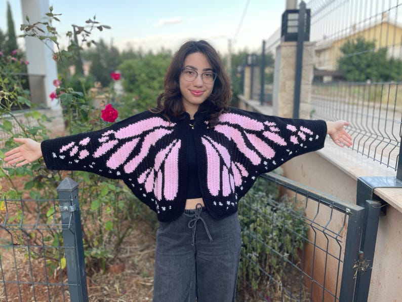 May include: A black crochet cardigan featuring a pink butterfly wing pattern on the sleeves and back. The garment has an open front design with wide, wing-like sleeves that extend outward, showcasing a detailed pink and black color block motif.