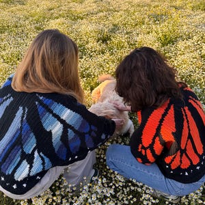 May include: Two people wearing black crochet butterfly wing sweaters, one with blue wings and the other with orange wings, are kneeling in a field of white flowers. The sweaters have detailed wing patterns. The scene is outdoors, with a dog in the center.