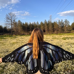 May include: A black crochet cardigan featuring a large butterfly wing pattern on the back. The wings are designed with shades of blue and light blue yarn, accented with small white spots along the edges. The garment has long sleeves and a wide fit.