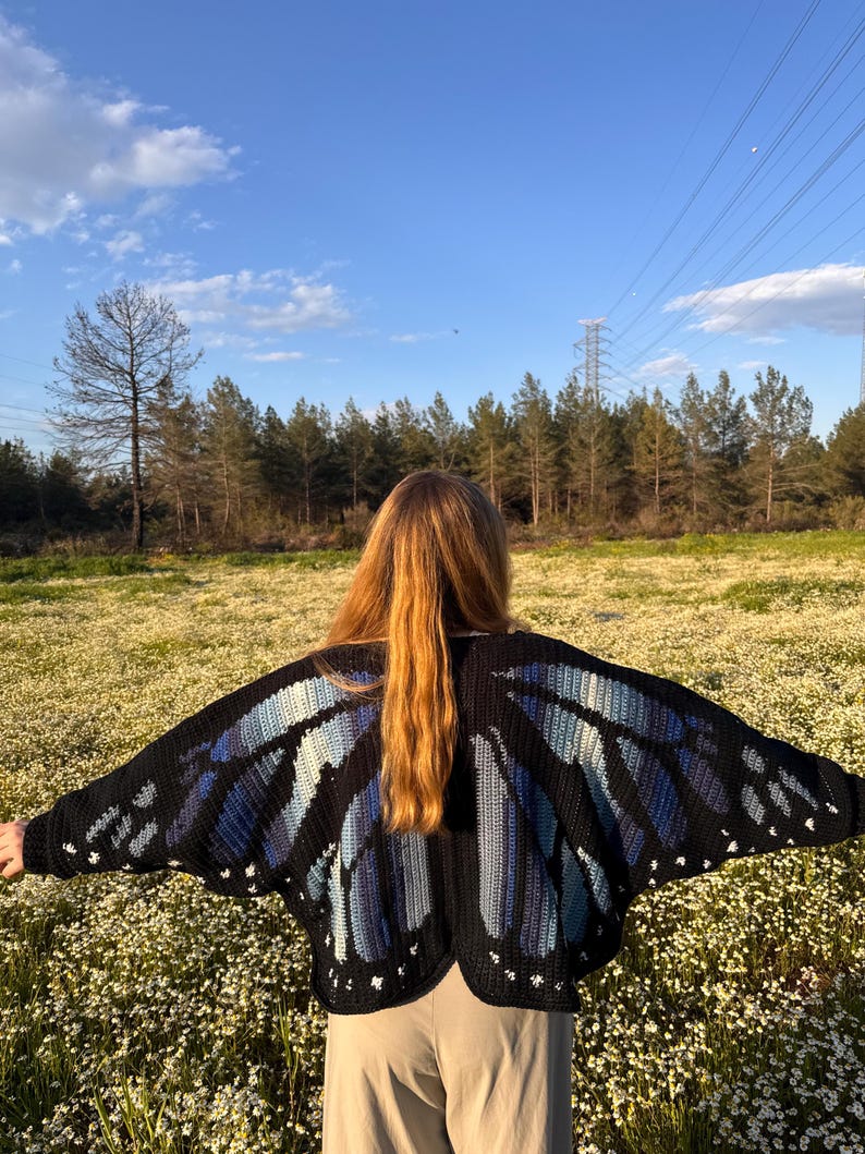 May include: A black crochet cardigan featuring a large butterfly wing pattern on the back. The wings are designed with shades of blue and light blue yarn, accented with small white spots along the edges. The garment has long sleeves and a wide fit.