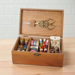 May include: A wooden sewing box with an open lid, revealing compartments filled with sewing supplies. The lid holds ornate gold scissors. Inside are jars of beads, colorful tapes, small paint bottles, and a floral pouch.