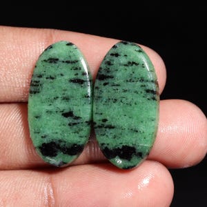 Può includere: Due pietre preziose di zoisite di forma ovale. Le pietre hanno un vibrante colore di base verde con inclusioni e segni neri. Le pietre preziose sono lucidate e hanno una superficie liscia e lucida.