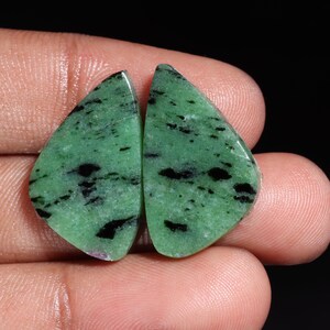 May include: Two polished gemstones with a triangular shape. The stones are a vibrant green color with black inclusions and a smooth, glossy surface. The gemstones are held in a hand against a black background.