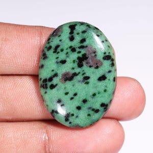 May include: Oval-shaped polished gemstone with a green base color and black and maroon inclusions. The stone has a smooth, glossy surface and is held in a hand. The gemstone is likely used for jewelry or decorative purposes.
