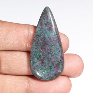 May include: A teardrop-shaped gemstone with a polished surface. The stone features a mottled pattern of deep red and teal green hues. The gemstone is held in a hand, showcasing its size and shape. This could be used for jewelry making.
