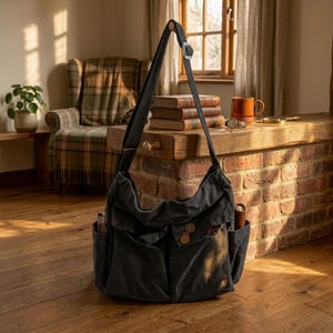 Puede incluir: Un bolso mensajero de lona gris oscuro con una correa larga ajustable. El bolso tiene múltiples bolsillos, incluidos dos bolsillos delanteros con cierres de botón de madera y dos bolsillos laterales. El bolso está colgado en una habitación con suelo de madera.