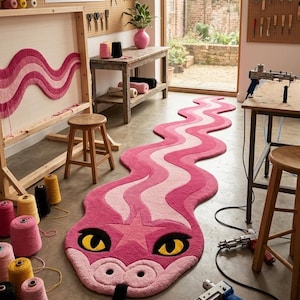 Puede incluir: Una alfombra larga y ondulada con forma de serpiente en tonos rosa y rosa claro, con una estrella en la cabeza, ojos amarillos y una lengua negra. La alfombra se exhibe sobre un suelo de hormigón en un taller, rodeada de ovillos, taburetes y pistolas de tufting.
