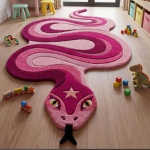 Könnte beinhalten: Ein großer, schlangenförmiger Teppich mit einem Farbverlauf in Rosa- und Magentatönen. Der Teppich hat einen stilisierten Kopf mit einem fünfzackigen Stern auf der Stirn, gelben Augen und einer schwarzen gespaltenen Zunge. Der Körper windet sich.