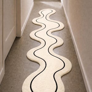 Puede incluir: Una alfombra ondulada de color crema con un borde negro, colocada en un pasillo. El diseño de la alfombra se asemeja a un río que fluye o a una forma abstracta. La alfombra está hecha de un material suave y texturizado.