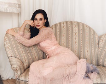 May include: A woman wearing a light pink, long-sleeved dress with a lace overlay, sitting on a patterned sofa. The dress has a fitted bodice and a flowing skirt. The sofa has a floral and striped pattern. The woman has dark hair.
