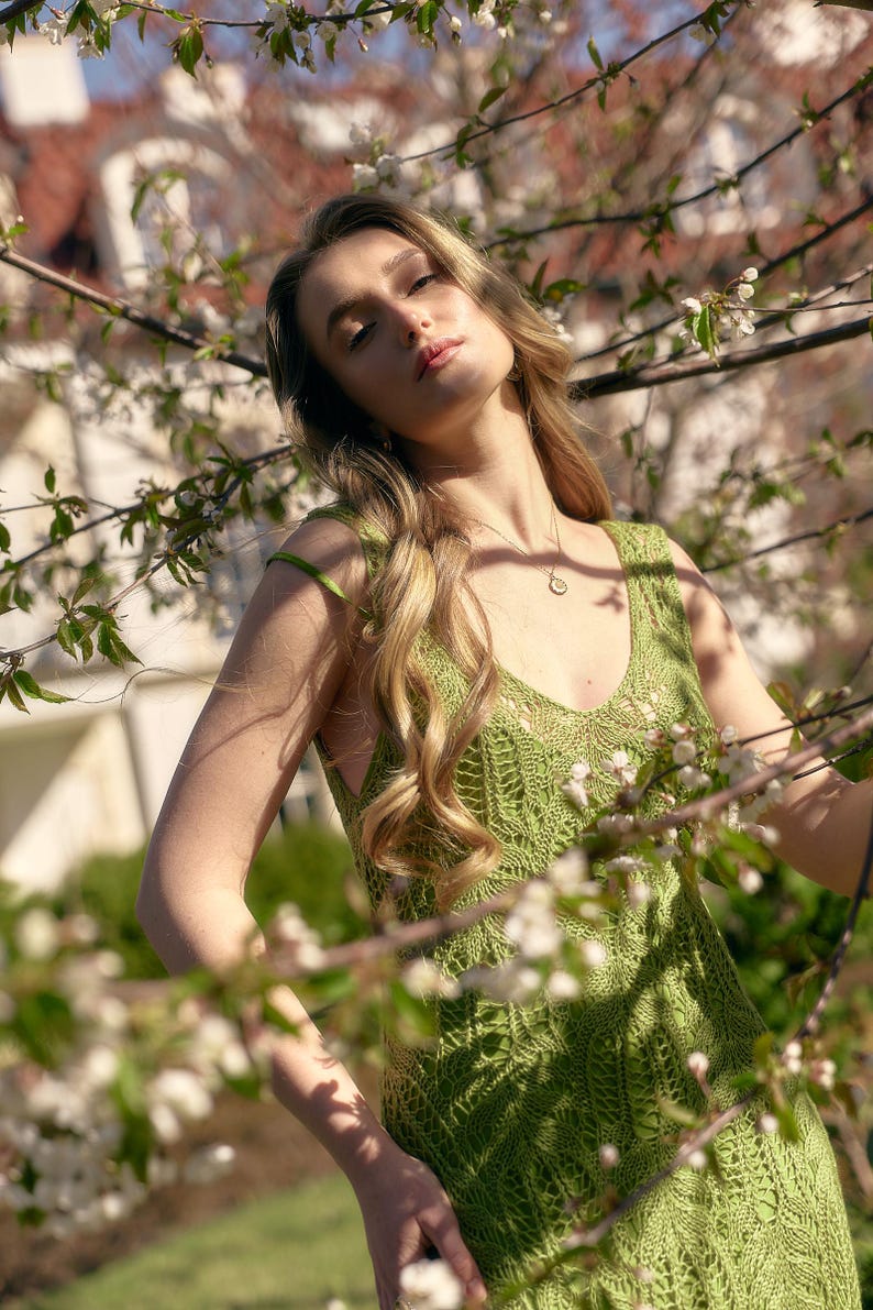 May include: A light green, sleeveless, crocheted dress with a V-neck. The dress is worn outdoors, surrounded by white flowering branches. A gold necklace is visible.