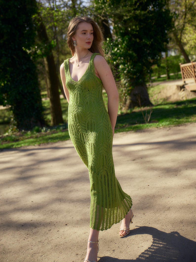 May include: A sleeveless, green, knitted dress with a V-neck. The dress has a detailed leaf pattern on the bodice and a flowing skirt. The model is wearing gold-colored sandals. The background is a park with trees and sunlight.