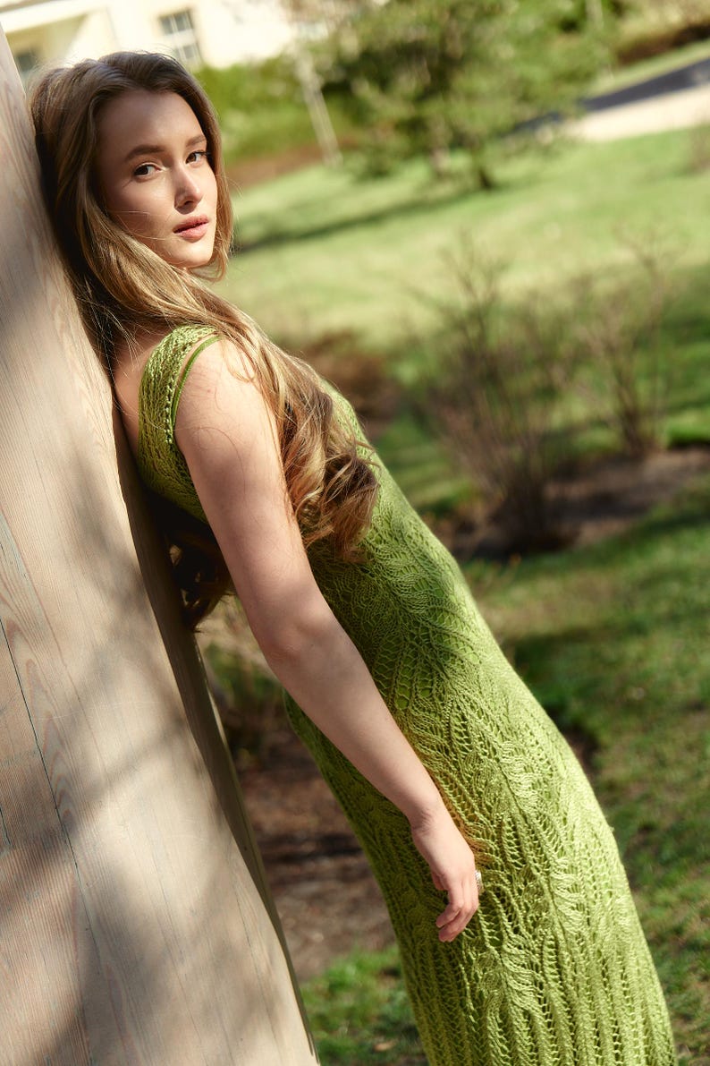 May include: A sleeveless, light green dress with a detailed, open-weave pattern. The dress is worn outdoors, with the wearer leaning against a wooden structure. The background features green grass and trees, suggesting a sunny day.