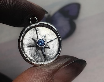 May include: A silver pendant featuring a compass rose design with a central blue gemstone. The circular pendant has a textured edge and a loop for a chain. The background is blurred, with a hint of a butterfly.