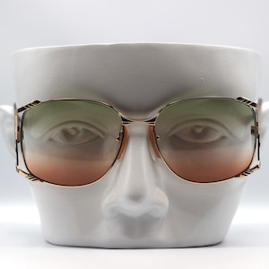 May include: Gold-framed sunglasses with gradient lenses, displayed on a white ceramic face sculpture. The lenses transition from green to orange. The sunglasses have a square shape and a classic design.