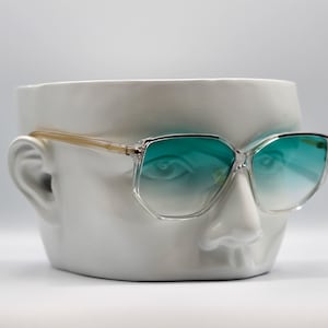 Puede incluir: Gafas de sol con montura transparente y lentes degradadas turquesas sobre una cabeza de cerámica blanca. Las gafas tienen forma cuadrada con una patilla dorada. Las lentes hacen una transición de turquesa a transparente.