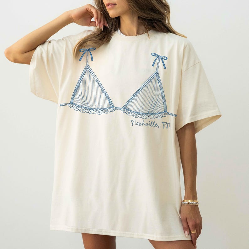 Pode incluir: Camiseta creme com um desenho a tra&ccedil;o azul de um suti&atilde; com listras verticais e acabamento em renda. O design do suti&atilde; inclui pequenos la&ccedil;os nas al&ccedil;as. O texto "Nashville, TN" est&aacute; impresso abaixo.