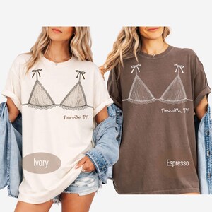 Pode incluir: Duas camisetas, uma marfim e outra espresso, cada uma com um desenho a tra&ccedil;o preto de um suti&atilde; com detalhes de la&ccedil;o. O texto "Nashville, TN" &eacute; impresso abaixo do desenho do suti&atilde;. A camisa marfim tem a palavra "Ivory" impressa, e a camisa espresso tem a palavra "Espresso" impressa.