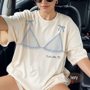 Puede incluir: Camiseta color marfil con un dibujo lineal azul de un sujetador con lazos y encaje. El texto "Nashville, TN" está impreso debajo. La camiseta tiene un corte holgado.