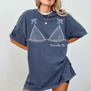 Pode incluir: T-shirt cinza escuro com um desenho branco de suti&atilde; com la&ccedil;os e renda. O texto "Nashville, TN" est&aacute; impresso abaixo do desenho do suti&atilde;. A camiseta tem mangas curtas e um corte folgado.