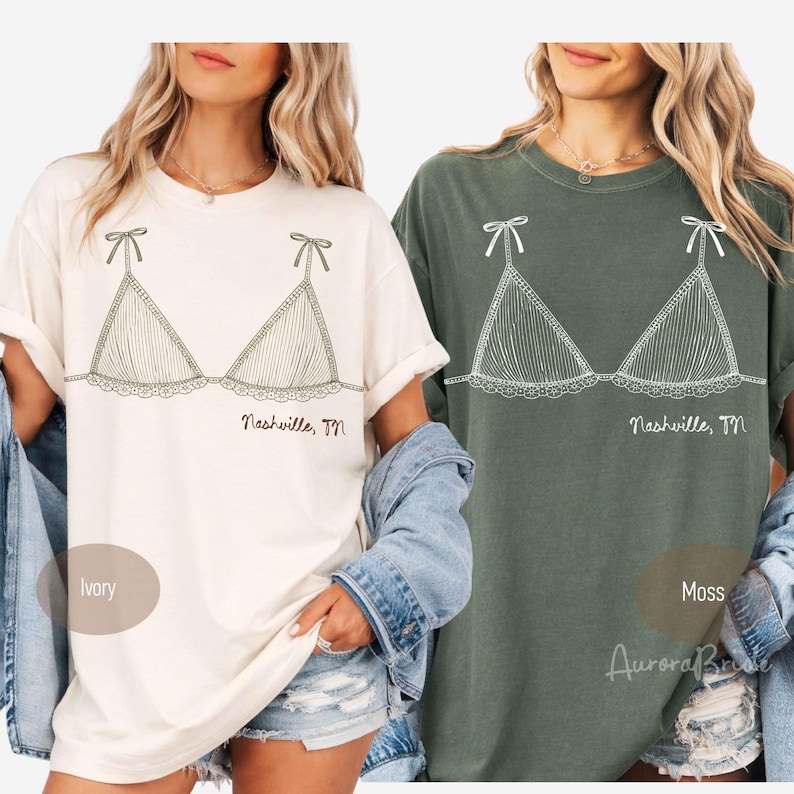 Pode incluir: Duas camisetas, uma marfim e outra verde musgo, cada uma com um desenho a tra&ccedil;o branco de um suti&atilde;. O design do suti&atilde; inclui la&ccedil;os, renda e linhas verticais. O texto "Nashville, TN" est&aacute; abaixo. As etiquetas "Ivory" e "Moss" est&atilde;o presentes.
