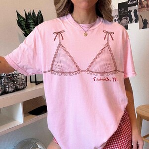 Pode incluir: T-shirt rosa claro com um desenho a tra&ccedil;o vermelho de um suti&atilde; com la&ccedil;os e detalhes de renda. O texto "Nashville, TN" est&aacute; impresso abaixo. A camiseta tem mangas curtas.