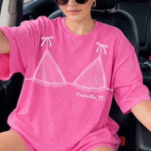Pode incluir: Uma t-shirt rosa brilhante com um desenho a tra&ccedil;o branco de um soutien com la&ccedil;os nas al&ccedil;as. O texto "Nashville, TN" est&aacute; impresso abaixo do desenho do soutien. A t-shirt &eacute; de estilo casual e oversized.