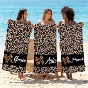 May include: Three beach towels with a leopard print pattern. Each towel features a black band with a bow and a name in white script: Grace, Aria, and Hannah. The towels are held by three people on a beach.