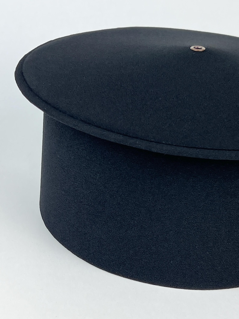 May include: A black hat with a rounded top and a cylindrical base. The hat has a small, circular metal detail on the top. The hat's design is simple and classic, suitable for various occasions.