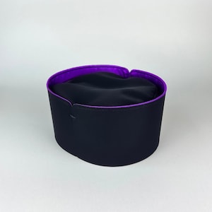 May include: A black hat with a purple interior lining. The hat has a cylindrical shape with a flat top and a small opening at the front. The hat is likely made of a smooth fabric.